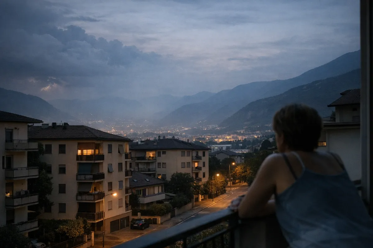 Persona affacciata a un balcone con vista su palazzi e luci della città nella valle, tra foschia e nubi serali